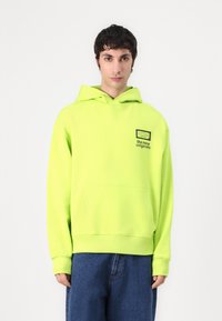 Helle grüne Kapuzenjacke aus weichem Stoff, mit einer Kängurutasche und einem schwarzen Logo-Patch auf der Brust. Lässige, oversized Passform.