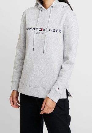 Personne portant un sweat à capuche gris clair Tommy Hilfiger avec logo et texte "EST. 1985", associé à un pantalon noir sur un fond uni.