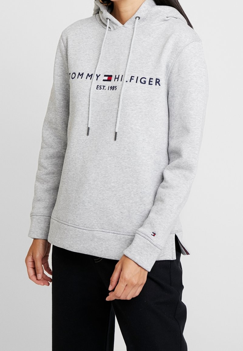 Personne portant un sweat à capuche gris clair Tommy Hilfiger avec logo et texte "EST. 1985", associé à un pantalon noir sur un fond uni.
