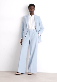 Costume bleu clair sur mesure avec des pantalons à jambes larges et un blazer structuré, associé à une chemise blanche à col et des chaussures beiges. Texture de tissu lisse.