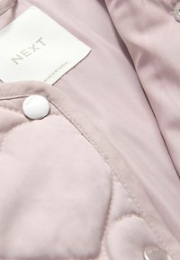 Chaqueta acolchada de color rosa claro con una textura suave, botones de presión plateados y una etiqueta blanca que indica "NEXT, HECHO EN MYANMAR."