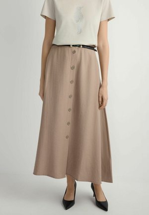 Kobieta ubrana w beżową midi spódnicę z przodu na guziki, białą koszulkę z wzorem ptaka, czarny pasek i czarne szpiczaste wysokie obcasy, stojąca na białej podłodze.