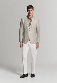 Homme portant un blazer beige, une chemise grise, un pantalon blanc et des chaussures noires, debout devant un fond uni gris clair.