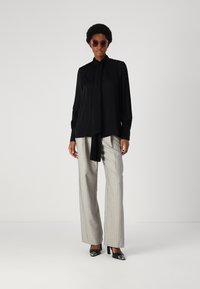 Schwarze Bluse mit langen Ärmeln und Bindedetail, kombiniert mit hellgrauen gestreiften Hosen und schwarzen Absätzen. Das Model trägt übergroße Sonnenbrillen.
