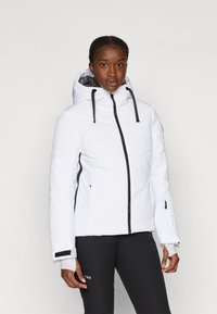 Fehér pufferszövet kabát, geometrikus steppelésű mintával, fekete cipzárral, állítható kapucnival és mandzsettákkal, fekete nadrág felett viselve.