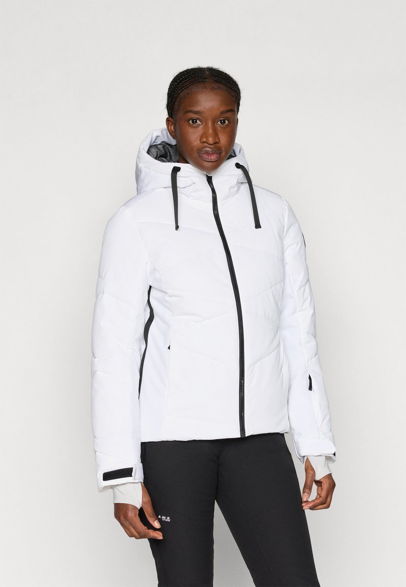 Fehér pufferszövet kabát, geometrikus steppelésű mintával, fekete cipzárral, állítható kapucnival és mandzsettákkal, fekete nadrág felett viselve.