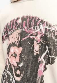Sudadera beige con un estampado gráfico que presenta la cara de un león y elementos tormentosos en rosa y negro. El material parece suave y texturizado.