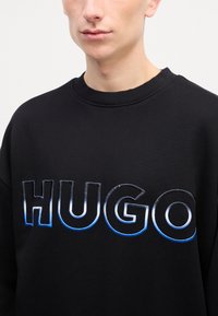 Felpa nera con un grande logo "HUGO" in lettere blu e bianche sfumate, con collo e polsini a costine, realizzata in tessuto morbido.