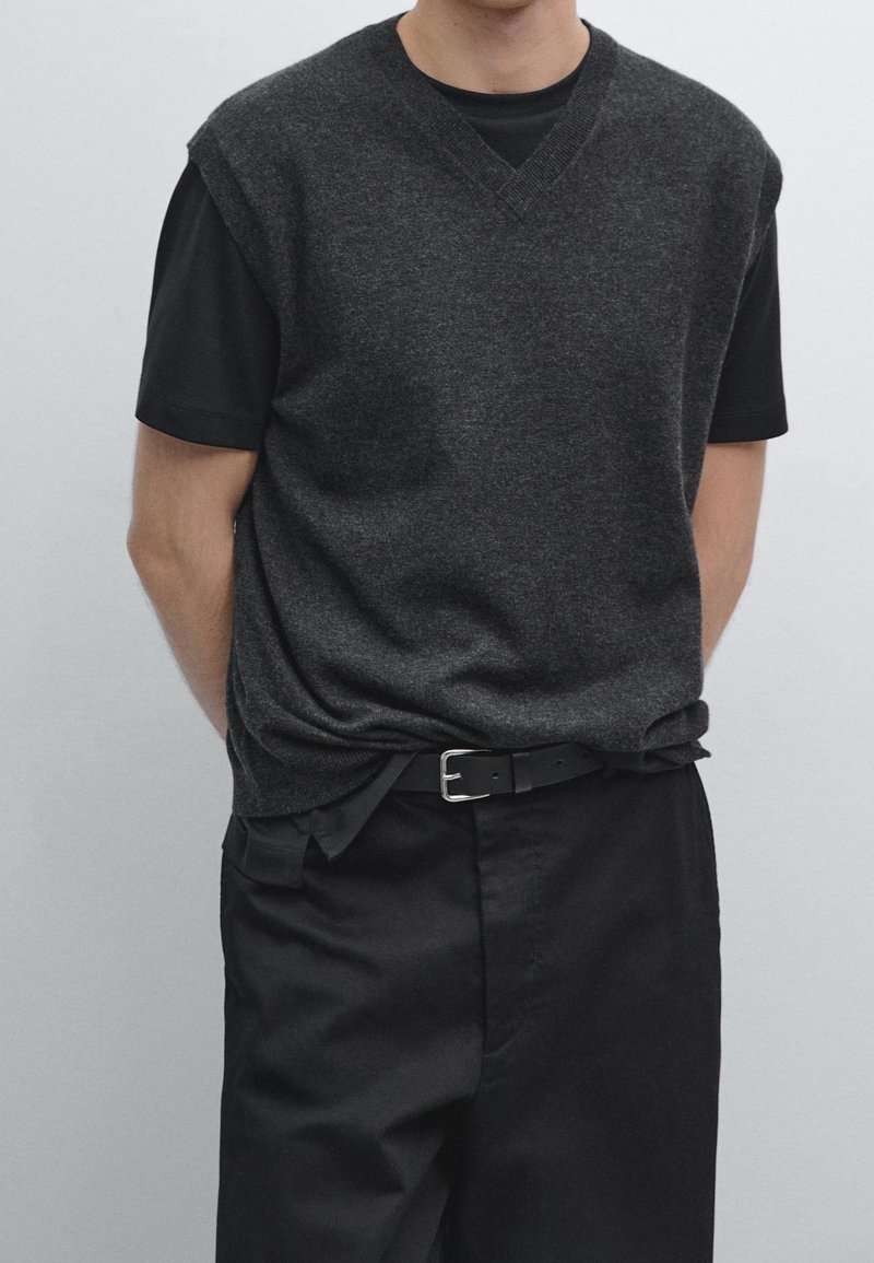 Personne portant un pull sans manches gris foncé par-dessus une chemise noire à manches courtes, un pantalon noir et une ceinture noire avec une boucle argentée.
