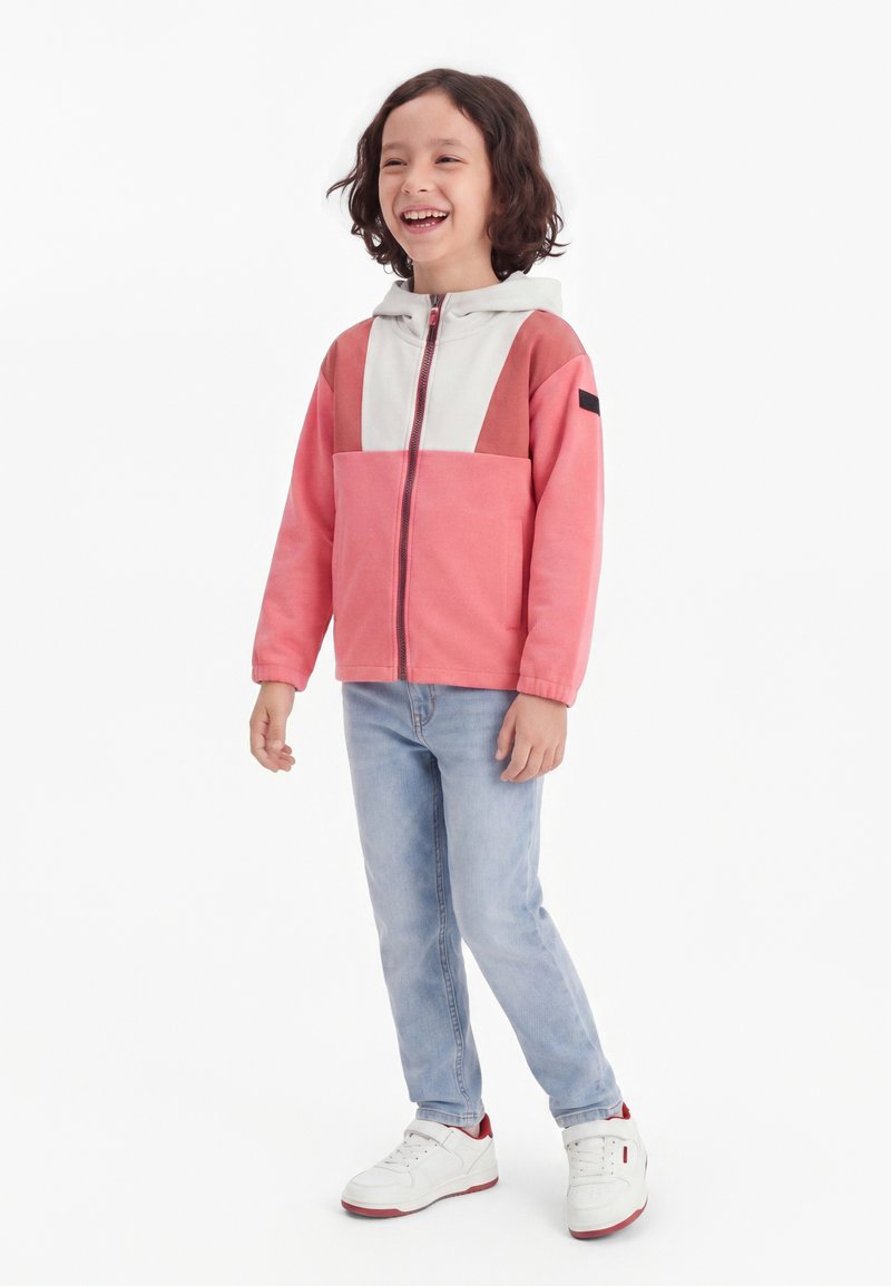 Lächelndes Kind mit lockigem Haar, das eine rosa-weiße Kapuzenjacke, hellblaue Jeans und weiße Sneakers mit roten Akzenten trägt.