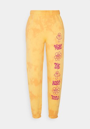 Pantalon de jogging tie-dye jaune avec des motifs roses de champignons souriants et de fleurs ainsi que le texte "Have the best time" sur la jambe droite.