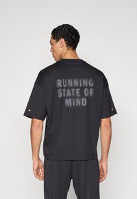 Černé tričko s krátkým rukávem a voln�ým střihem, s nápisem "RUNNING STATE OF MIND" vytištěným na zádech v bledém stylu.