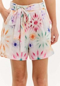 Žena v bielych bavlnených šortkách s farebnými vzormi tie-dye v tvare hviezdneho výbuchu a s tkaninovým opaskom zviazaným do mašle v páse.