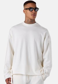 Homme portant un sweat-shirt blanc surdimensionné et un pantalon assorti, lunettes de soleil noires, tatouage au cou et petite boucle d'oreille, se tenant devant un fond uni.