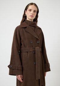 Brązowy zamszowy płaszcz trencz z dwurzędowym zapięciem, dużymi klapami i szerokimi mankietami. Zawiera dopasowany pasek i czarne guziki.