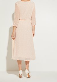 Hellrosa gepunktetes Kleid mit drei Viertelärmeln, tailliertem Schnitt und Knopfakzenten an den Bündchen, kombiniert mit weißen High-Heels.