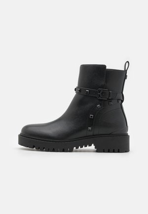 Botín de cuero negro hasta el tobillo con punta redondeada, material texturizado, hebilla decorativa, tachuelas cuadradas y suela robusta.