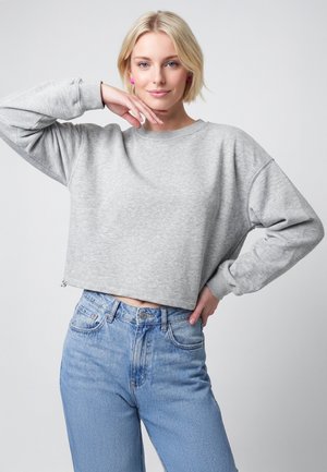 Ung kvinde med kort blondt hår iført en grå cropped sweatshirt og blå jeans, poserer med den ene hånd på hoften og den anden nær ansigtet.