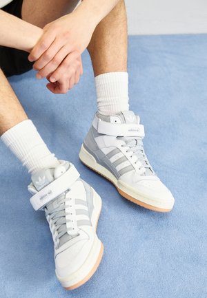 Personne portant des baskets montantes Adidas blanches et grises avec des chaussettes blanches côtelées, assise sur un tapis bleu clair, les mains reposant sur le genou.
