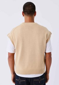 Jersey sin mangas de color beige con cuello y dobladillo acanalados, con un acabado texturizado, llevado sobre una camiseta blanca y unos vaqueros.