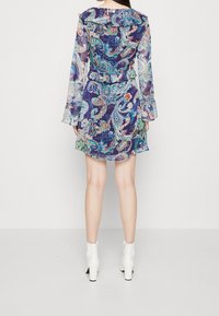 Fialové šaty s paisley vzorem, s průsvitnými dlouhými rukávy, rozšířenými manžetami, volánkovým výstřihem a rozšířeným okrajem, doplněné bílými kotníkovými botami.