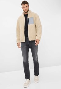 Giacca in pile beige con tasca pettorale grigia con cerniera, indossata sopra una maglietta nera e abbinata a jeans scuri e sneakers chiare.