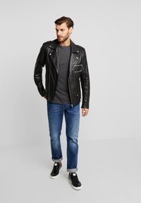 Svart skinnbikerjakke med teksturert overflate, sølvglidelåser, grå T-skjorte, blå jeans og svarte sneakers med hvite såler.