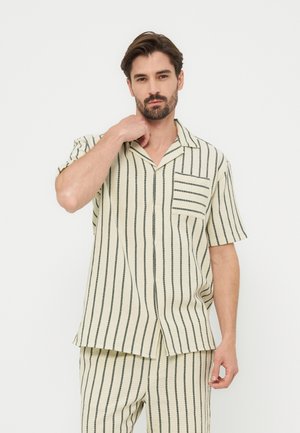 Άνδρας με γενειάδα, ντυμένος με κρεμ πουκάμισο και παντελόνι με κάθετες μαύρες ρίγες, ποζάρει με το ένα χέρι κοντά στον λαιμό, μπροστά σε απλό φόντο.