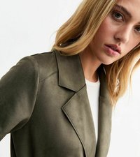 Blazer in suede verde oliva, con rever a scossese e vestibilità su misura. Cuciture visibili e texture liscia. Indossato sopra una maglietta bianca.