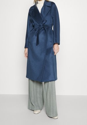 Femme portant un long manteau bleu marine ceinturé, un col roulé blanc, un pantalon large gris et des chaussures blanches sur un fond uni.