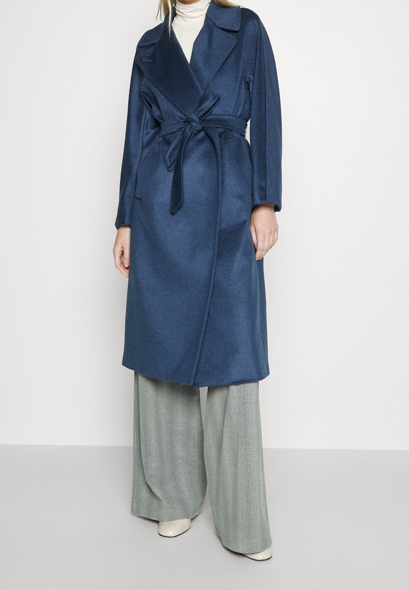 Femme portant un long manteau bleu marine ceinturé, un col roulé blanc, un pantalon large gris et des chaussures blanches sur un fond uni.