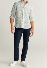 Mann trägt ein hellblaues, blumiges Kurzarmhemd mit aufgerollten Ärmeln, dunkelblaue Slim-Fit-Jeans und weiße Sneakers, steht vor einem schlichten Hintergrund.