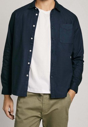 Man draagt een losgeknoopt donkerblauw overhemd over een wit T-shirt en kaki broek, met één hand in de zak, tegen een effen achtergrond.