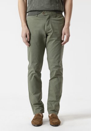 Hombre con pantalones de corte recto color verde oliva y mocasines de ante marrón, de pie frente a un fondo blanco liso.