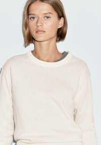 Ljust beige tröja med rund halsringning, mjuk textur, långa ärmar och en avslappnad passform, lager-på-lager över en mörkgrå topp.