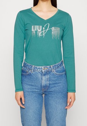 Vrouw draagt een teal lange mouwen V-hals shirt met zilveren "Liu Jo" tekst en lichtblauwe high-waist jeans, staand tegen een effen achtergrond.