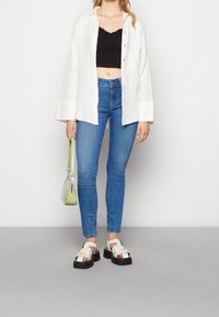 Kvinna i en svart crop top, öppen vit skjorta med knappar, blå skinny jeans och vita platåsandaler, som håller en liten grön handväska.