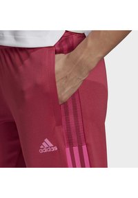 Różowe spodnie sportowe z teksturowanej tkaniny, z bocznymi kieszeniami i akcentami z siateczki, ozdobione trzema paskami oraz logo Adidas w jasnoróżowym kolorze.