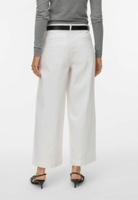 Persona que lleva pantalones blancos de pierna ancha, cinturón negro, camiseta gris de manga larga y sandalias negras con tacón, de pie contra un fondo claro y liso.