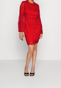 Röd spetsklänning med långa genomskinliga ärmar med klockform, scallopad nederkant och texturerad blommönster. Bärs med svarta klackar med öppen tå och en metallisk clutch.