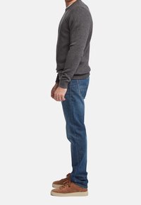 Homme debout de côté portant un pull gris, un jean bleu et des baskets marron sur un fond blanc uni.