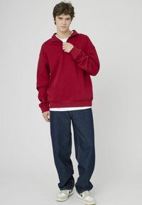 Pull-over en polaire rouge avec un col zippé au quart, associé à un jean en denim foncé à coupe décontractée et des chaussures de sport blanches et grises.