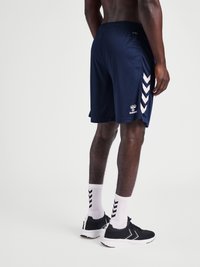 Mann som har på seg marineblå sportshorts med hvite chevron-striper, hvite sokker og svarte treningssko, står foran en ensfarget bakgrunn.