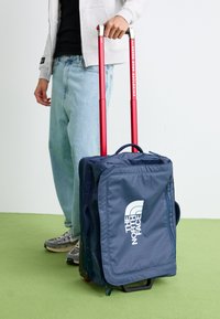 Valise à roulettes bleu marine avec une texture lisse, dotée d'une poignée extensible rouge, d'une poignée supérieure et d'un logo blanc sur le côté. Des sangles de rangement sont visibles.