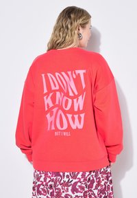 Sweatshirt corail à coupe oversize, avec un texte rose audacieux qui dit "JE NE SAIS PAS COMMENT MAIS JE VAIS LE FAIRE." Poignets et ourlet côtelés.