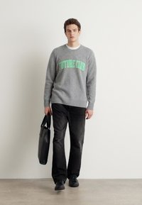 Grå genser med "FUTURE CLUB" i grønn tekst, kombinert med svarte jeans og sko. Holder en svart veske. Enkel, moderne design.
