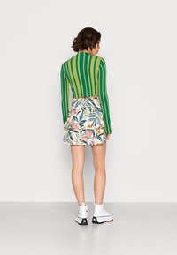 Top de manga longa às riscas verde e amarelo, saia branca com padrão floral rosa e sapatilhas altas brancas com detalhes pretos, vistas por trás.