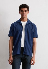 Jonge man met kort, krullend haar die een marineblauw overhemd met korte mouwen draagt over een wit T-shirt, tegen een effen witte muur.