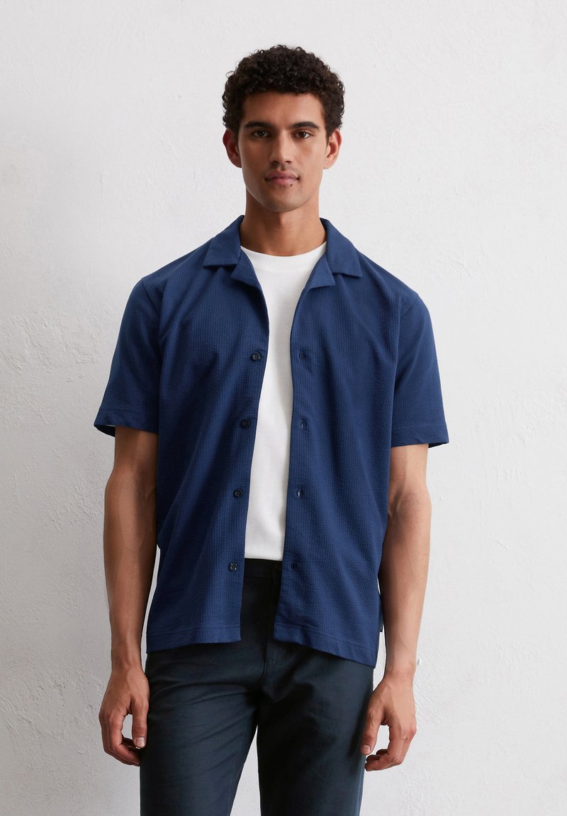 Jonge man met kort, krullend haar die een marineblauw overhemd met korte mouwen draagt over een wit T-shirt, tegen een effen witte muur.
