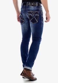 Jean en denim bleu foncé avec une coupe slim, présentant des coutures contrastées et des poches arrière à motifs uniques. Portés avec des chaussures en cuir marron.
